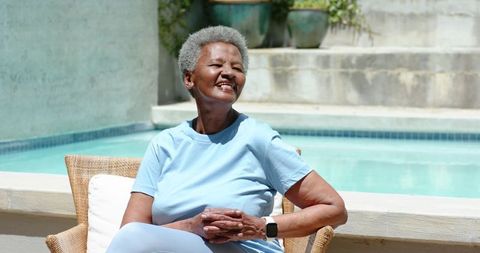 Senior african woman smiling and relaxing by pool wearing smartwatch and light blue outfit