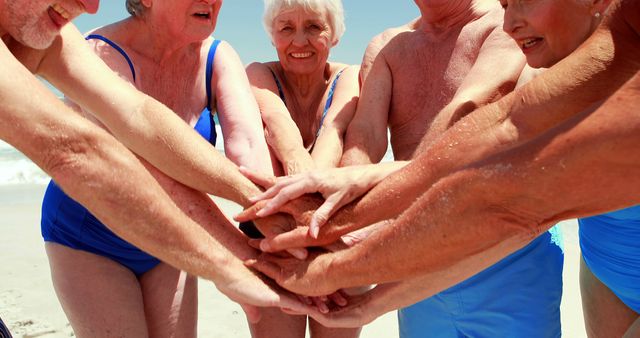 Seniors in swimwear standing at beach stacking hands in a show of solidarity and friendship. Useful for themes related to active aging, senior group activities, friendship, retirement lifestyle, beach vacations, and community support.