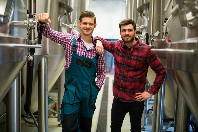 Two male brewers standing confidently in a brewery, surrounded by stainless steel tanks. One is wearing overalls, and the other is in a plaid shirt. Both are smiling and appear professional. Ideal for use in articles or advertisements related to the brewing industry, beer production, teamwork, and industrial work environments.