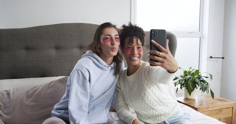 Two women taking selfie on bed wearing pink under-eye patches cozy morning routine