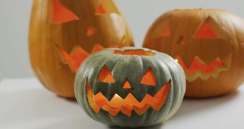 Glowing carved green-gray pumpkin with lit orange jack-o'-lanterns on white tabletop