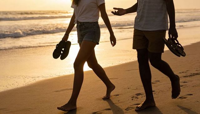 Couple walking barefoot on golden sunset beach holding sandals leaving footprints on shore