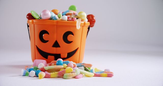 A Halloween-themed orange bucket filled with a variety of colorful candies spills its contents onto a surface, with copy space. The festive container suggests a celebration of the Halloween holiday, often associated with trick-or-treating and sweet treats.
