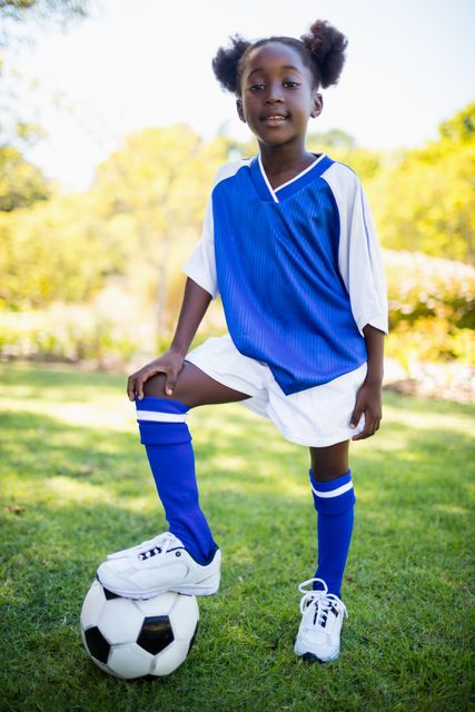 Young girl wearing blue and white soccer uniform standing with one foot on a soccer ball in a park. Ideal for use in advertisements for youth sports programs, athletic wear, outdoor activities, and children's health and fitness campaigns.