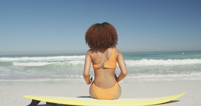 Woman with curly hair in an orange bikini sits on a yellow surfboard facing the ocean. Ideal for promoting beach holidays, summer vacations, surfing activities, and tropical getaways. Suitable for lifestyle and leisure advertising.