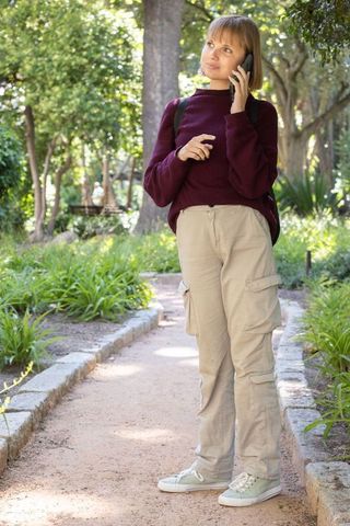 African american woman talking on phone in sunlit park path, casual outfit with backpack