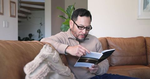 Mature asian man reading brochure comfortably on leather sofa at home