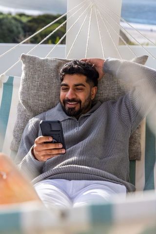 Relaxed man reclining in hammock chair on ocean-view balcony, scrolling smartphone