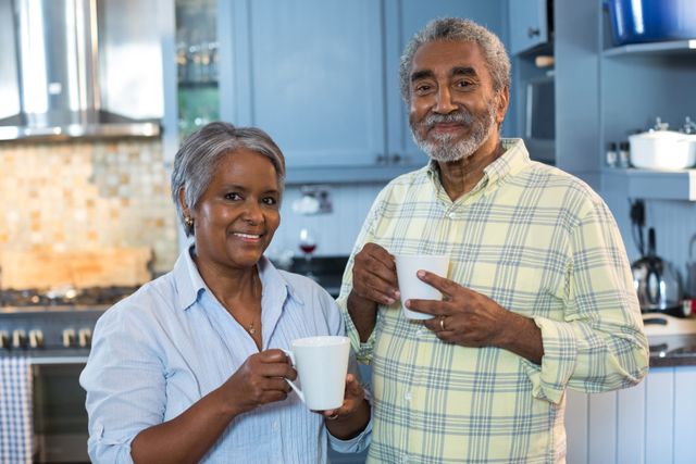 Senior couple standing in a modern kitchen, holding coffee cups and smiling. Ideal for use in advertisements, articles, and blogs about senior living, healthy lifestyles, and family moments.