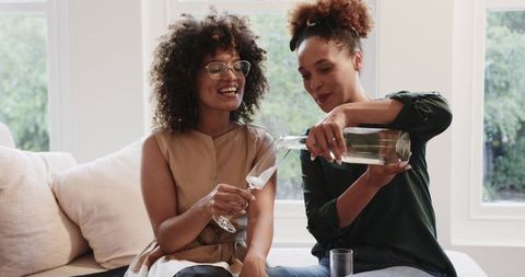 African American women pouring sparkling water and laughing on cozy sunlit sofa