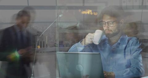 Bearded man working on laptop drinking coffee with city reflection on office window