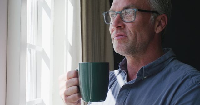 Happy caucasian man wearing glasses, looking through window and drinking coffee. Spending time at home alone.