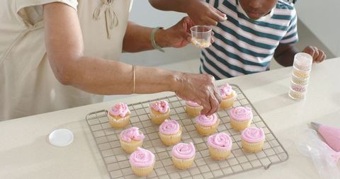 African grandmother and granddaughter decorating pink frosted cupcakes, intergenerational baking