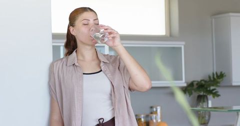 Woman drinking water in sunlit modern kitchen, wearing beige shirt and white tank top