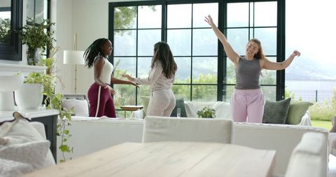 Joyful diverse women dancing and laughing in sunlit modern living room with large windows