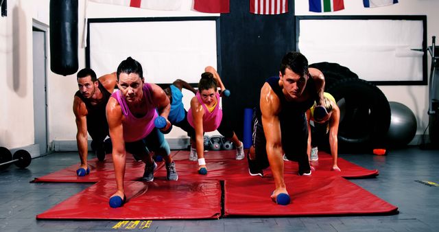 A group of people engaged in fitness training using dumbbells, lying on fitness mats in gym. They perform strength training exercises in a gym setting. This image can be used for advertisements promoting fitness centers, workout programs, health and wellness blogs, and personal training services.