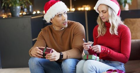 Couple tagging christmas gifts on sofa wearing santa hats and cozy holiday sweaters