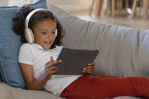 School-age girl reclining on couch using tablet with headphones for learning and play