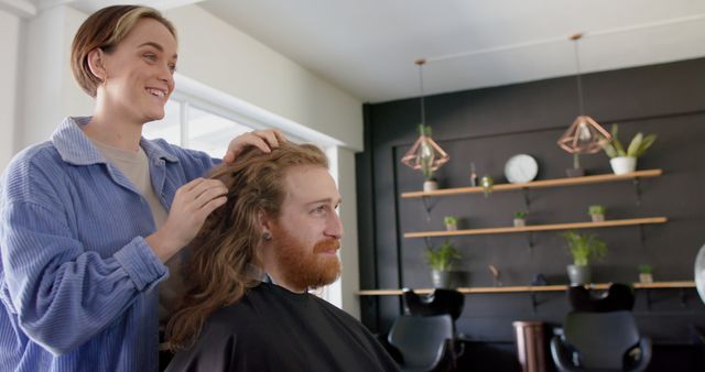 This depicts a female hairdresser styling a male client's long hair in a modern salon. The male client sits comfortably while the hairdresser works. The salon has a contemporary design with geometric light fixtures and shelves with plants. This can be used for content related to hairdressing services, customer satisfaction, modern salons, hairstyling tips, and professional hair care environments.