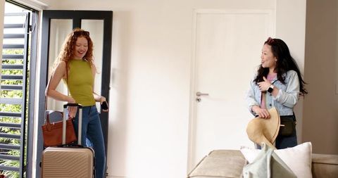 Redhead and asian woman greeting during home arrival with rolling suitcase and straw hat