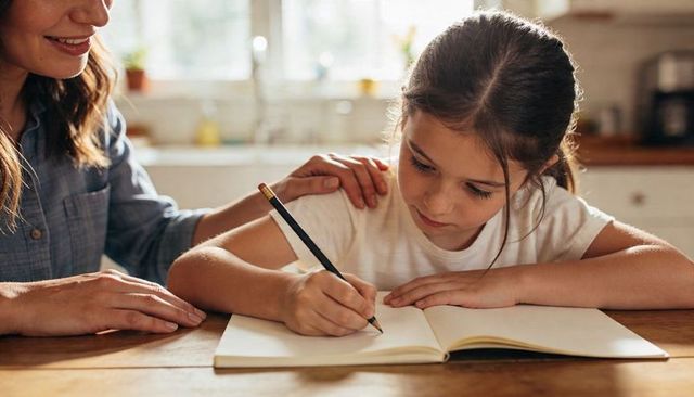 Young girl writing in notebook at sunlit kitchen table with supportive parent