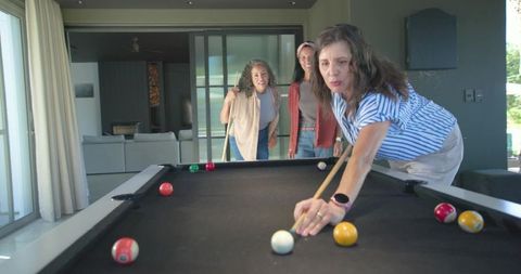 Middle-aged women playing pool at home, leaning woman lining up shot on black-felt table