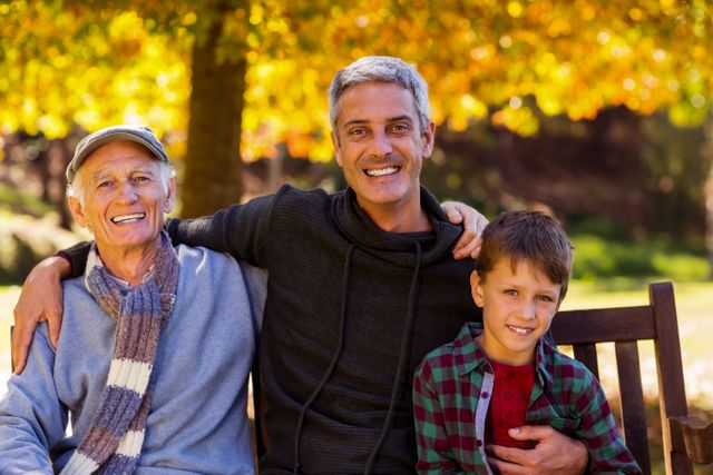 Three generations of men sitting on a bench in a park during autumn. The grandfather, father, and son are smiling and enjoying the beautiful fall colors. This image is perfect for illustrating family bonding, generational relationships, and outdoor activities in nature. It can be used in advertisements, family-oriented articles, and promotional materials for parks or outdoor events.