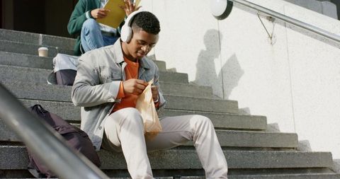African american student sitting on campus steps listening to music opening lunch bag