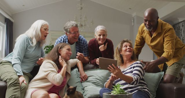 Group of senior friends gathered in a living room, enjoying social time around a digital tablet. Ideal for use in articles about senior living, technology adoption by the elderly, or promoting social connectivity among older adults. Can also be used in marketing materials targeting senior-specific products or services.