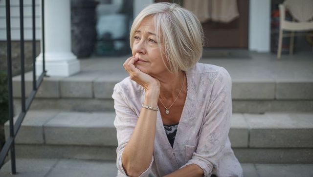 Thoughtful senior woman sitting and leaning on front porch steps wearing pink blouse