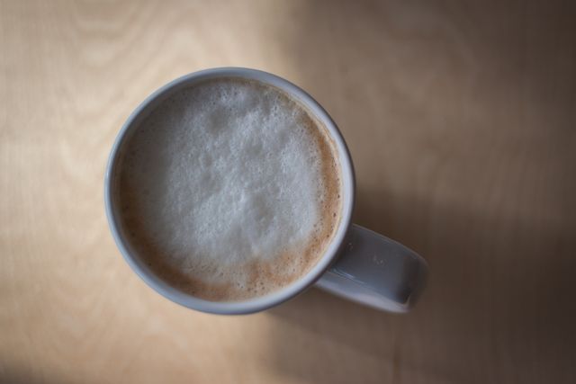 A warm and inviting top view of a frothy cappuccino in a white mug, placed on a wooden table. The froth on the coffee highlights its creamy texture, making it appear freshly made and tempting. The warm lighting contributes to a cozy atmosphere, making it suitable for use in content related to coffee culture, cafés, morning routines, breakfast imagery, and articles or posts about relaxation and enjoying beverages.