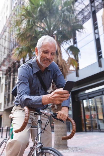 Senior man riding a bike in the city while using a smartphone and smiling. Ideal for promoting active lifestyles, urban living, technology use among seniors, and outdoor activities. Suitable for advertisements, blog posts, and articles related to health, fitness, and modern technology.