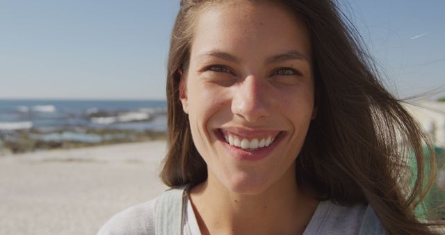Young woman smiling at camera while enjoying a sunny day at the beach. Background features ocean and sandy shore, indicating a relaxed and joyful moment. The image is perfect for marketing materials related to travel, relaxation, vacation, or lifestyle promotions.