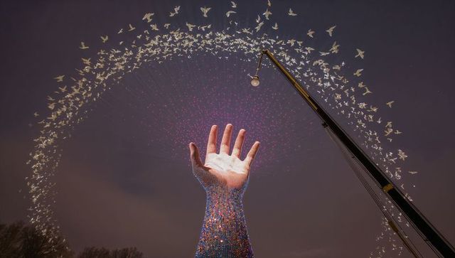 Giant led hand sculpture reaching sky at night radiating pink light with arcing bird flock