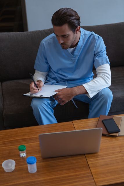 Male surgeon in blue medical uniform writing on clipboard while sitting on sofa in hospital lobby. Suitable for use in healthcare, medical, and hospital-related content. Ideal for illustrating concepts of patient care, medical documentation, and healthcare professionals at work.
