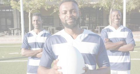 Confident rugby captain holding ball with teammates standing behind on sunny grass field