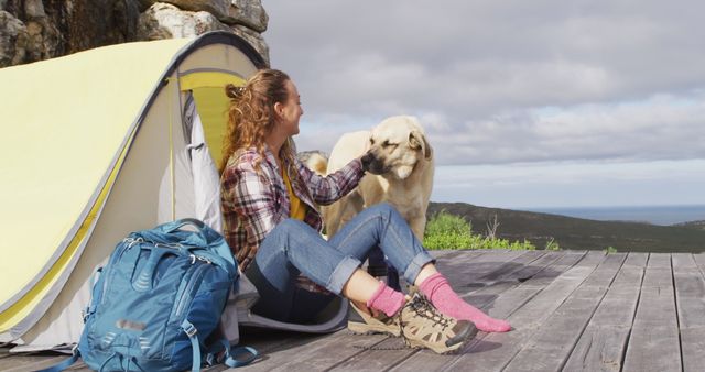 Woman sits in front of yellow tent on wooden deck with mountains and sea in background, patting a tan dog. Surrounded by hiking gear including backpack. Captures essence of adventure and outdoor lifestyle, emphasizing companionship with pet. Ideal for travel blogs, pet care promotions, and adventure tourism advertisements.