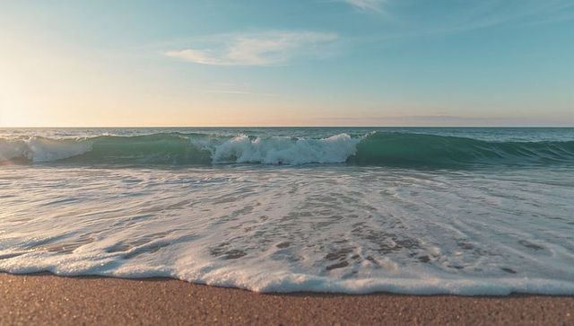 Breaking teal wave rolling onto wet sandy beach at sunrise with pastel dawn sky and white foam foreg
