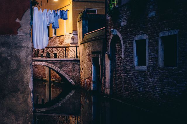 Quiet Venetian alley illuminated at night, featuring a small bridge over a water canal with hanging laundry. This can be used for themes related to travel, history, European cities, and urbanscapes.