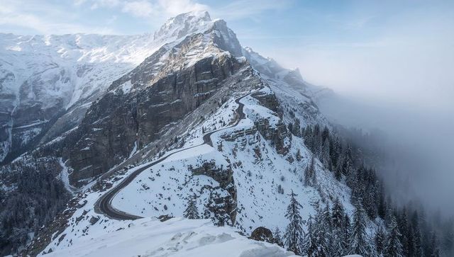 Winding alpine ridge road climbing toward snow-capped peak in misty winter morning