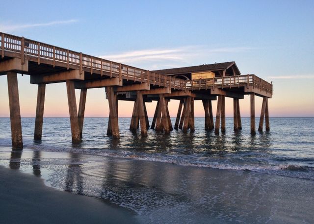 Wooden pier extending over the ocean with calm waters and clear skies during sunset. Gentle waves lap against the shore, reflecting soft evening light. Ideal for beach vacation promos, travel websites, relaxation themes, coastal living blogs, and inspiring social media content focusing on tranquility and natural beauty.