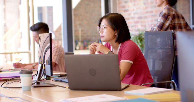 Young woman intently analyzing data on her computer in modern office setting with colleagues in background. Useful for themes related to office work, technology, data analysis, teamwork, professional life, and contemporary workspaces. Can be used in corporate websites, presentations, blog posts, and marketing materials showcasing productive work environments and teamwork.