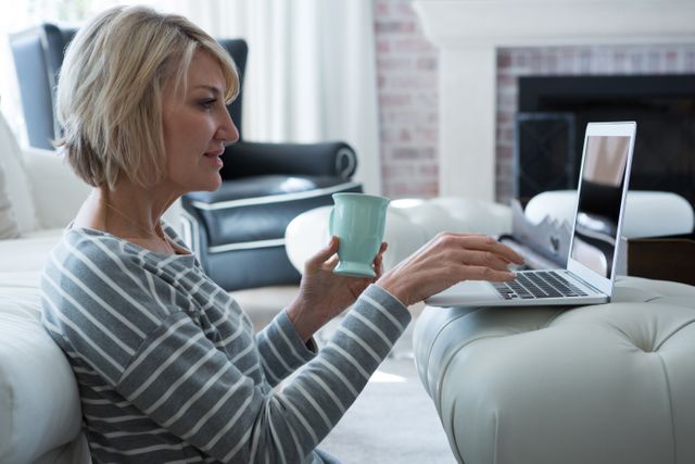 Mature woman sitting on floor in living room, using laptop and holding coffee mug. Ideal for concepts related to remote work, home office, relaxation, modern lifestyle, and technology use at home.