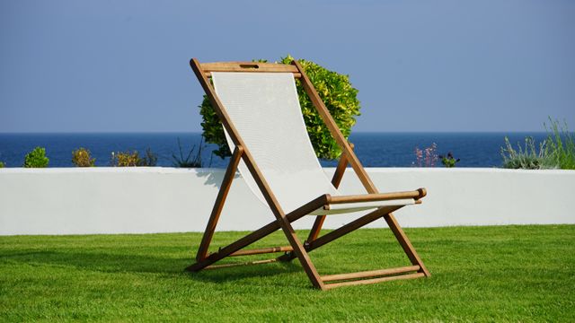 Wooden deck chair positioned on green lawn with clear view of ocean in background. Invokes feelings of relaxation and tranquility perfect for landscaping ideas, summer travel brochures, outdoor living promotions, and seaside vacation advertisements.
