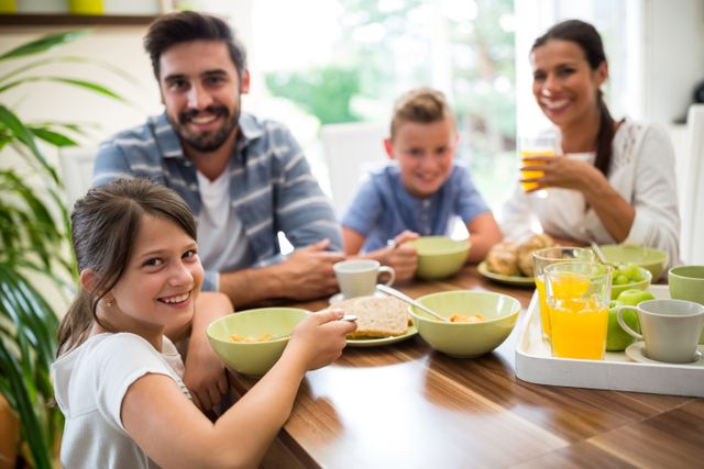 Family sitting around a table enjoying breakfast together. Includes children and parents with various breakfast items such as orange juice, cereal, and toast. Useful for illustrating family bonding, healthy eating, morning routines, and happy family time.