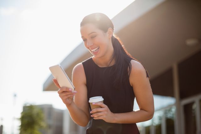 Businesswoman standing outside a conference center, using a mobile phone and holding a coffee cup. She is smiling, indicating a positive interaction or successful communication. Ideal for use in business, technology, and professional networking contexts, as well as for promoting corporate events, modern work environments, and urban lifestyles.