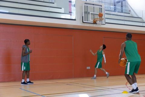 Multicultural teen teammates practicing basketball on indoor hardwood court shooting layup in green