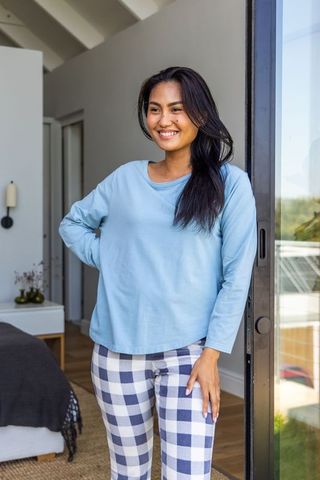 Asian woman smiling in doorway wearing blue top and checkered pajama pants, cozy bedroom