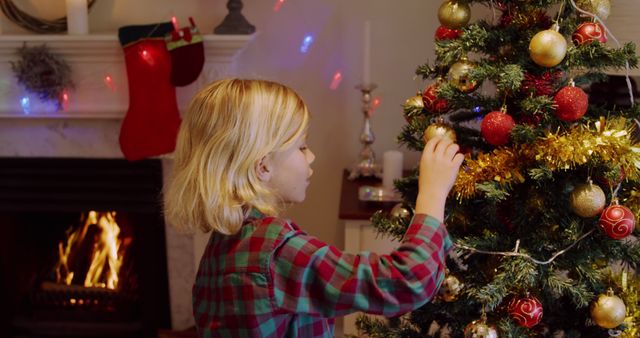 Image depicts a child decorating a Christmas tree by a fireplace. Ideal for use in holiday-themed promotions, festive greeting cards, adverts for winter celebrations, or content wishing holiday joy.