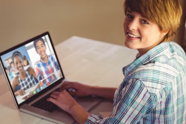 Teen looking at camera while using laptop for video call with friends, smiling. Suitable for concepts of online communication, digital interactions among teenagers, or remote learning/education. Can be utilized in advertisements for telecommunication services or educational tools.